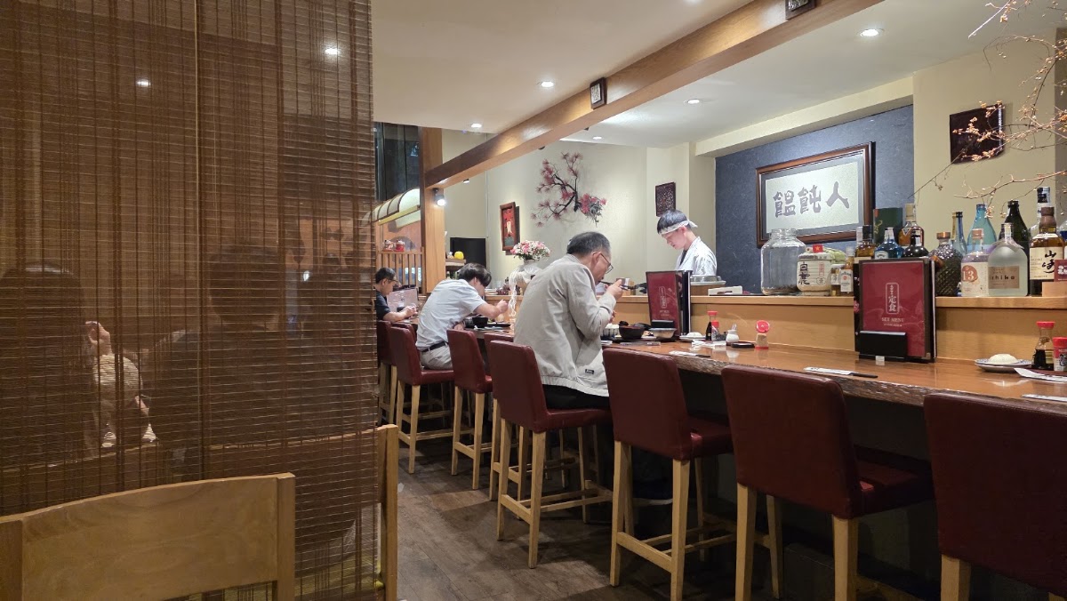 Quầy gỗ dài với ghế cao bọc nệm đỏ burgundy, thực khách đang ăn, đầu bếp phía sau quầy