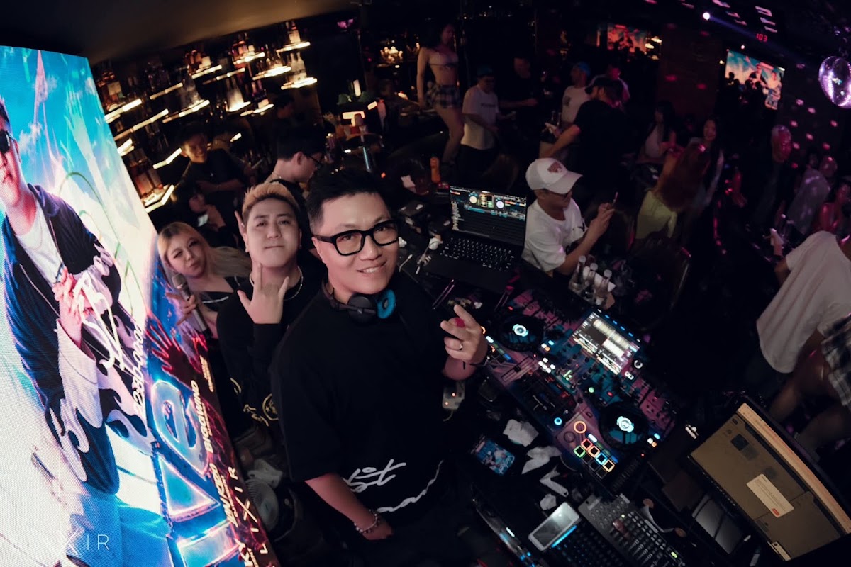 Ảnh chụp tại khu DJ: DJ đeo tai nghe, laptop, hình ảnh LED nhiều màu và đám đông xung quanh