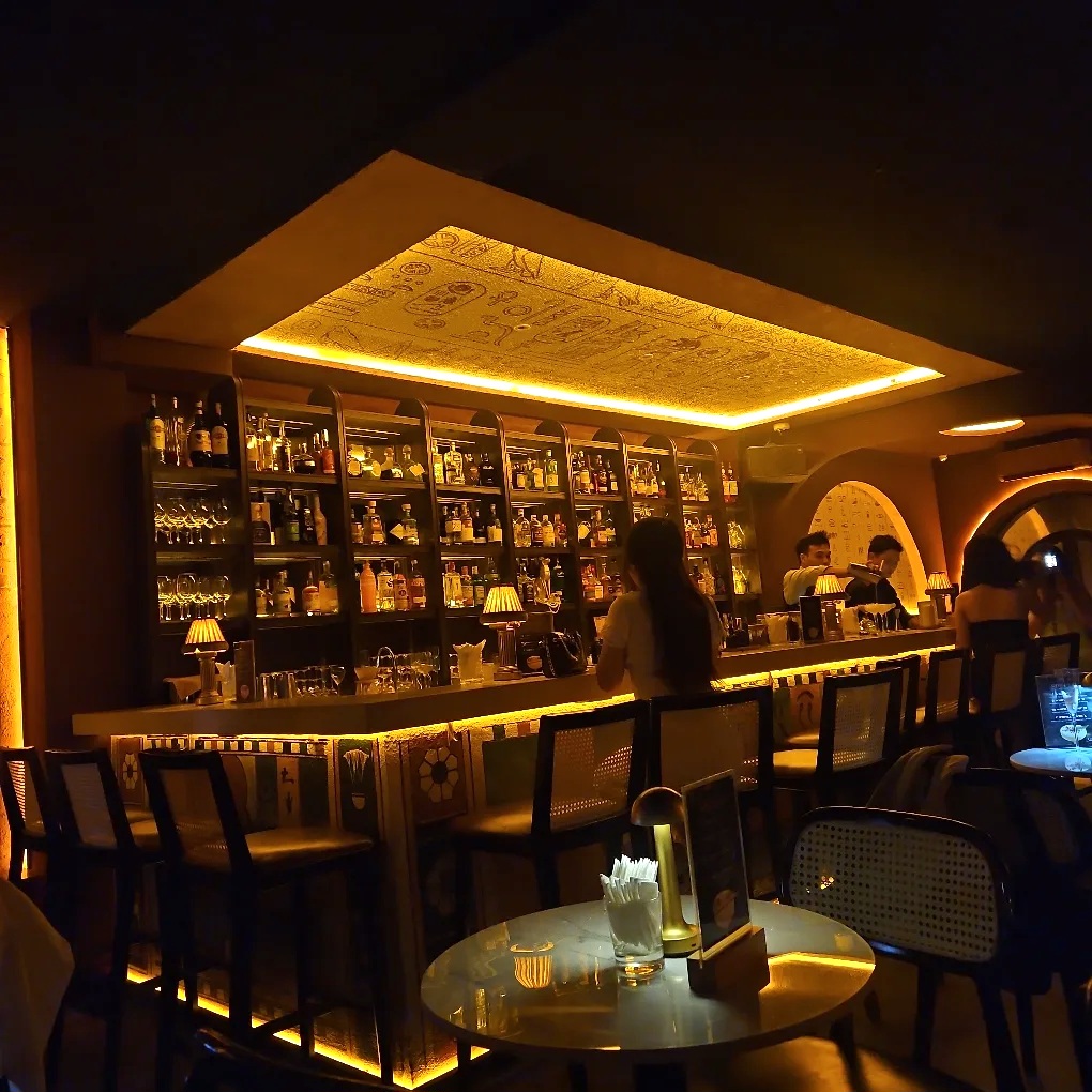 Interior wide — long bar counter, rattan stools, golden hieroglyph ceiling panel, backlit bottle wall