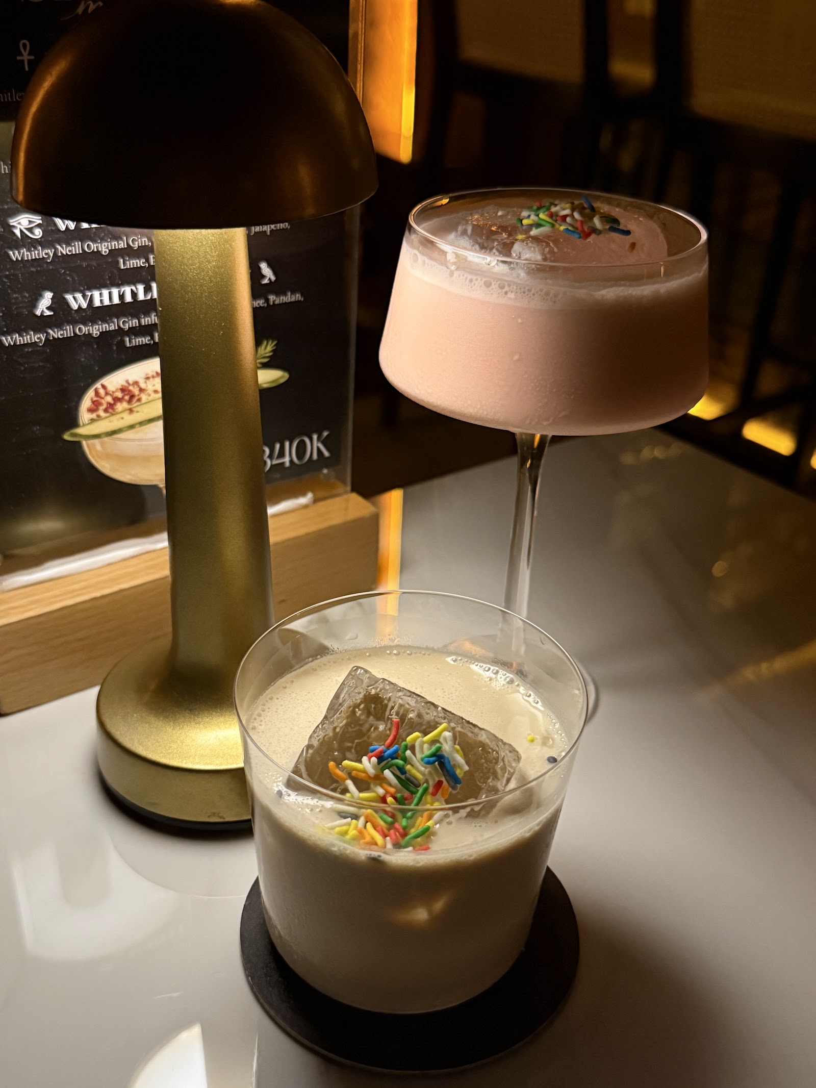 Two sprinkle-topped cocktails beside brass dome table lamp and Whitley Neill gin menu card (340K)