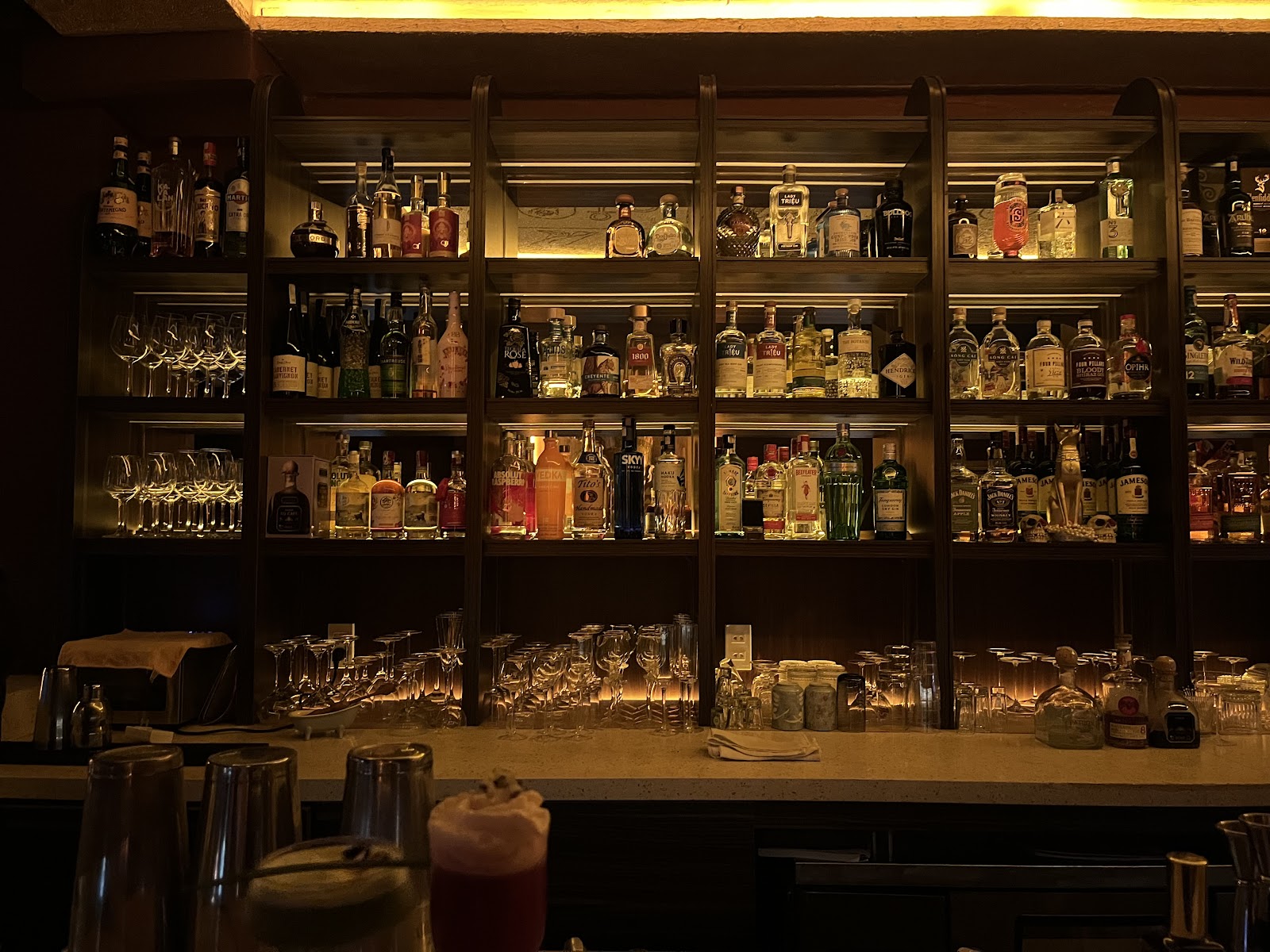Cận cảnh quầy bar sau — chai phát sáng, ly, dụng cụ pha chế