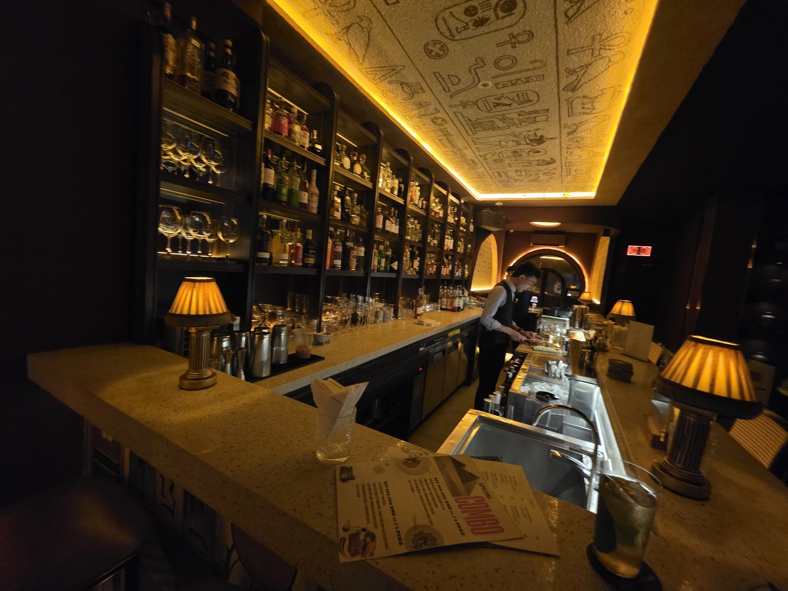 Along the bar counter — bartender at work, pleated-shade lamps, open paper menu booklet, hieroglyph ceiling
