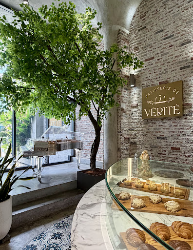 360° interior wide: indoor tree, brick + brass sign, marble counter with croissants, glass KAWAI piano, patterned tile