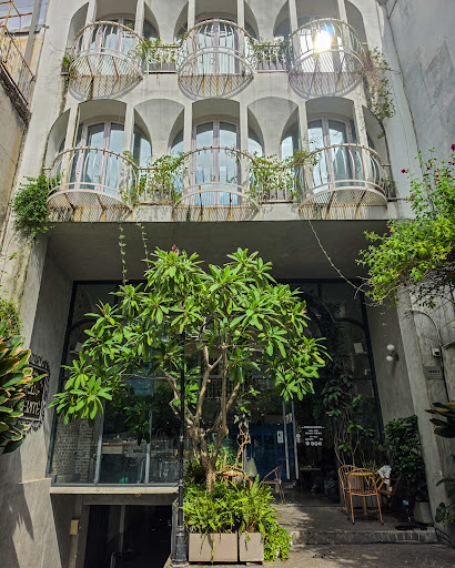 Storefront: tall narrow shophouse, arched windows, iron balconies + trailing plants, glass ground floor + hanging sign, frangipani tree, rattan chairs